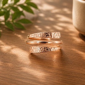 Peut inclure: Une bague bypass couleur or rose au design multicouche. La bague se compose de trois anneaux distincts : deux anneaux extérieurs avec une finition martelée et texturée, et un anneau central lisse et poli qui les relie en spirale.