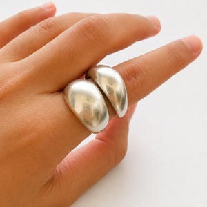 Peut inclure: Une bague argentée composée de deux segments lisses, arrondis et en forme de dôme qui entourent le doigt. Le métal présente une surface polie et réfléchissante avec un design incurvé et épais qui crée un aspect audacieux et continu.