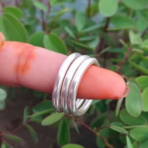 May include: A set of three polished silver-toned rings with a smooth, rounded band design. The rings are stacked together, featuring a reflective metallic surface that catches the light, showcasing a simple and uniform circular shape.
