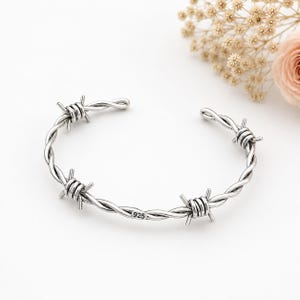 May include: A silver cuff bracelet designed to resemble twisted barbed wire with three distinct wire-wrapped spikes along the band. The metal surface features a small, clear stamp reading 925, indicating the sterling silver composition of the piece.