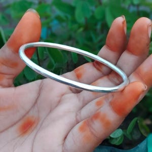 Puede incluir: Una pulsera tipo brazalete de tono plateado, lisa y pulida, con un diseño circular simple y continuo. El brazalete presenta un perfil redondeado y una superficie metálica reflectante, mostrando una estética limpia sin adornos adicionales.