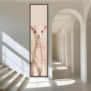 Könnte beinhalten: Ein vertikaler, rechteckiger Wandkunstdruck, der ein flauschiges weißes Lamm mit großen dunklen Augen und rosigen Wangen zeigt. Das Lamm trägt ein zartes, hellrosa Band, das zu einer Schleife um den Hals gebunden ist, vor einem neutralen cremefarbenen Hintergrund.
