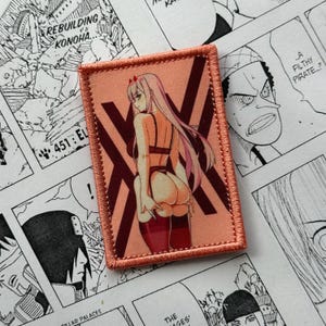 Può includere: Una toppa rettangolare ricamata raffigurante un personaggio con lunghi capelli rosa, che indossa biancheria intima rosso scuro e calze autoreggenti. Il personaggio è ritratto di schiena su uno sfondo color pesca con accenti a forma di X.