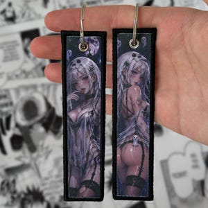 Puede incluir: Dos llaveros rectangulares de tela con ilustraciones estilo anime de un personaje con cabello blanco y ropa con temática de calaveras. Los diseños incluyen un fondo oscuro y fantasmal con una luna llena y pequeñas figuras espectrales con anillas.