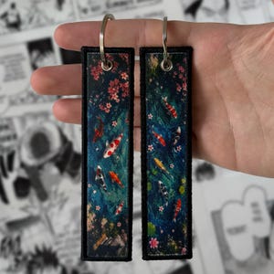 May include: Two rectangular fabric keychains featuring a dark blue water scene with colourful koi fish and pink cherry blossoms. Each keychain has a black stitched border and a silver metal split ring attached through a reinforced metal eyelet.