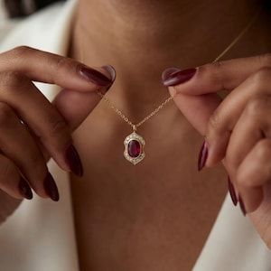 May include: A gold necklace featuring an oval-shaped red gemstone pendant surrounded by a halo of small white diamonds. The pendant has a decorative scalloped metal frame and hangs from a delicate gold chain, resting against the collarbone area.