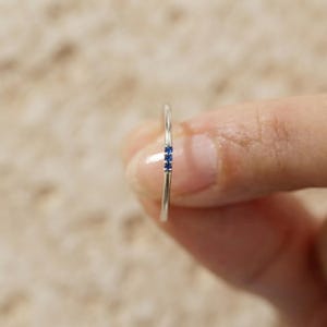 May include: A thin silver band ring featuring three small, round, deep blue gemstones set vertically in a row along the top of the band. The metal has a polished, reflective finish, and the stones are evenly spaced and securely mounted in place.