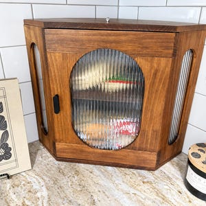 May include: A corner-shaped wooden storage cabinet with a medium brown finish. The cabinet features a central oval door and two side panels, all inset with vertical reeded glass. A small black handle is attached to the left side of the door.