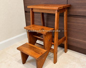 May include: A medium-brown wooden step stool with three tiers, featuring a sturdy frame and a rectangular top step. The text HandmadeWoodenvibe is printed in white across the middle step of this functional furniture piece.