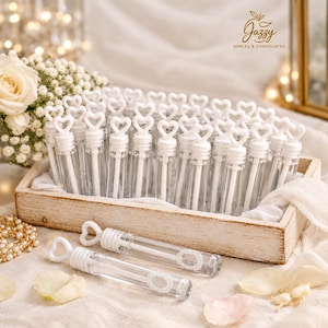 May include: A collection of clear plastic bubble wands with white heart-shaped handles, arranged in a rectangular wooden tray. Two additional bubble wands lie horizontally in front of the tray. The logo Jazzy Apples & Chocolates is visible.