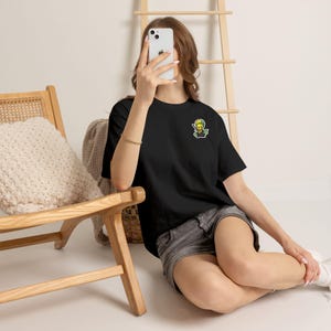 Może przedstawiać: Czarny t-shirt z krótkim rękawem, ozdobiony małą, kolorową grafiką przedstawiającą postać z kolczastymi włosami na lewej piersi. Koszulka ma okrągły dekolt i standardowy krój, wykonana z jednolitego czarnego materiału.