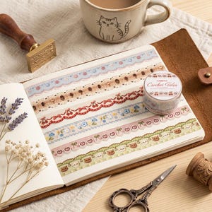 Könnte beinhalten: Ein offenes Notizbuch zeigt fünf Streifen dekoratives Klebeband mit Mustern aus Spitze, Kirschen, Schleifen, Blumen und kleinen Bären. Eine Rolle Klebeband mit der Aufschrift Crochet Tales liegt neben einer Keramiktasse mit Katzenmotiv, einem Stempel und einer Schere.