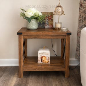 May include: A rectangular wooden end table with a dark brown finish, featuring black metal corner accents and an X-shaped side support. The top holds a light green textured vase with white flowers and a lamp. A box of Almond Panettone sits below.