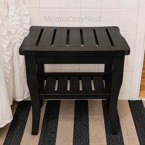 May include: A black wooden shower bench featuring a slatted top and a lower slatted shelf. The bench has four curved legs and the text MoonaCozyNest is visible on the wall behind it. The design includes a rectangular seat with open spaces.