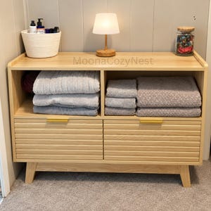 May include: A light wood storage console with two open shelves and two drawers featuring horizontal slats and gold handles. The top surface holds a white woven basket with bottles, a small lamp, and a glass jar. The text MoonaCozyNest is visible.
