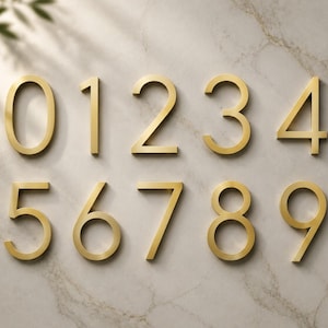 May include: A set of ten individual house numbers from zero to nine, crafted from a metallic gold material with a smooth finish. Each digit features a clean, sans-serif font style, arranged in two horizontal rows of five numbers each.