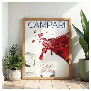 Può includere: Una stampa murale rettangolare incorniciata che presenta il testo CAMPARI e SODA con un'illustrazione grafica di un bicchiere che versa un liquido rosso intenso, accompagnato da cubetti di ghiaccio fluttuanti su sfondo bianco, in cornice di legno chiaro.