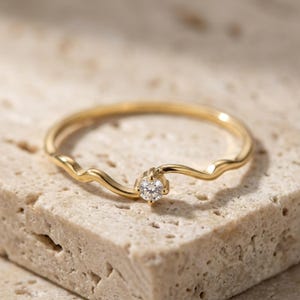 May include: A thin gold ring featuring a delicate, wavy band design. A single, small, round, clear gemstone is set in the centre of the band, held in place by a four-prong setting. The metal has a polished, reflective finish throughout the band.