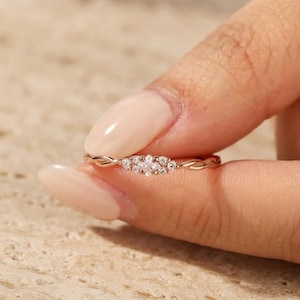 May include: A rose gold ring featuring a twisted band design with a central cluster of seven small, clear, round-cut stones arranged in a floral pattern. The metal band has a polished finish and a delicate, intertwined texture along the shank.