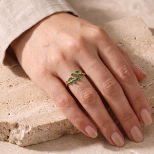 May include: A gold-toned ring featuring a delicate branch design adorned with small, marquise-cut green stones. The ring has an open-ended structure, with two leaf-shaped segments extending across the finger, showcasing vibrant green accents.