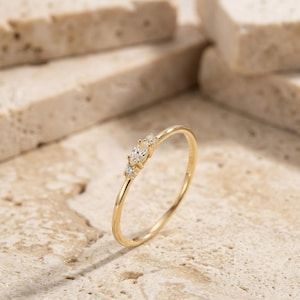 May include: A thin gold ring featuring a central marquise-cut clear stone flanked by two smaller round-cut clear stones. The band is smooth and polished, showcasing a delicate design with three stones set horizontally along the top of the ring.