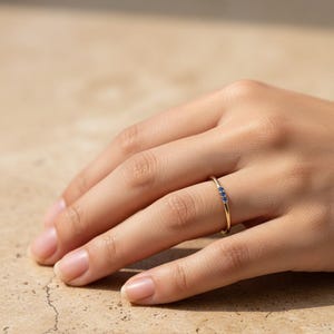 May include: A thin gold band ring featuring three small, round, vibrant blue gemstones set in a horizontal row. The metal has a polished finish, and the stones are evenly spaced along the top portion of the slender, circular ring band.