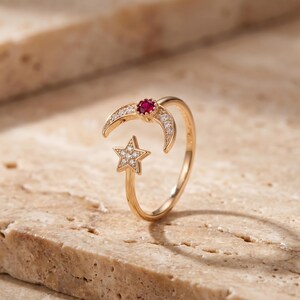 May include: A gold-toned open ring featuring a crescent moon motif adorned with small clear stones and a central round red gemstone, paired with a five-pointed star motif encrusted with clear stones at the opposite end of the band.