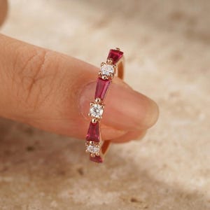 Peut inclure: Une bague en or rose ornée d'un motif alterné de rubis rouges taille baguette et de diamants blancs taille brillant ronde. Les pierres sont serties en ligne le long de l'anneau métallique, formant un design continu et symétrique.
