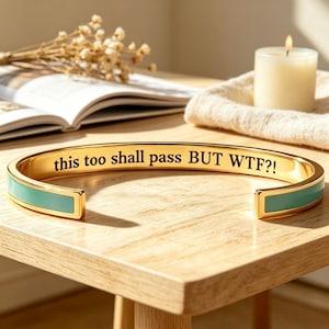 May include: A gold-toned cuff bracelet featuring a smooth, light teal enamel inlay along the outer band. The interior surface of the bracelet is engraved with the black text, this too shall pass BUT WTF?!, in a clear, legible serif typeface.