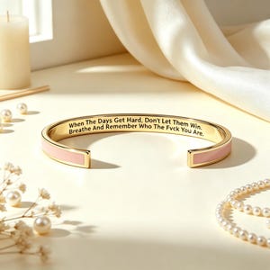 May include: A gold-tone cuff bracelet with a light pink enamel inlay on the exterior. The interior is engraved with the black text: When The Days Get Hard, Don't Let Them Win. Breathe And Remember Who The Fvck You Are. The bracelet is open-ended.