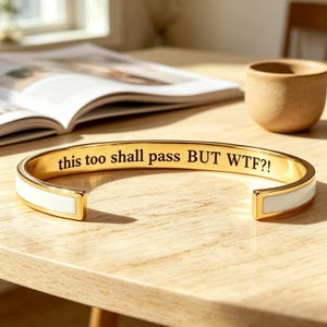 Puede incluir: Un brazalete dorado con una incrustación de esmalte blanco a lo largo de la banda exterior. La superficie interior está grabada con el texto negro "this too shall pass BUT WTF?!" en una fuente clara sin remates, creando contraste.