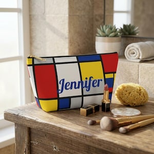 May include: A rectangular cosmetic bag featuring a geometric grid pattern in primary colors of red, yellow, and blue with black lines. The name Jennifer is printed in blue script across the center. The bag has a gold-toned zipper closure.