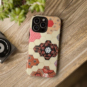 Puede incluir: Una funda para smartphone con fondo color crema y un patrón hexagonal repetitivo. El diseño incluye varios motivos florales y geométricos en tonos rojo, negro, rosa y azul, dispuestos en un estilo de patchwork acolchado.
