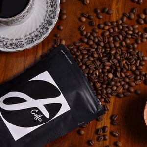 K&ouml;nnte beinhalten: Eine schwarze Kaffeet&uuml;te mit einem wei&szlig;en quadratischen Etikett, das einen stilisierten Buchstaben S und den Text Coffee zeigt, umgeben von verstreuten ger&ouml;steten Kaffeebohnen, einer wei&szlig;en Tasse mit schwarzem Blumenmuster und einem kleinen Holzsch&auml;lchen.