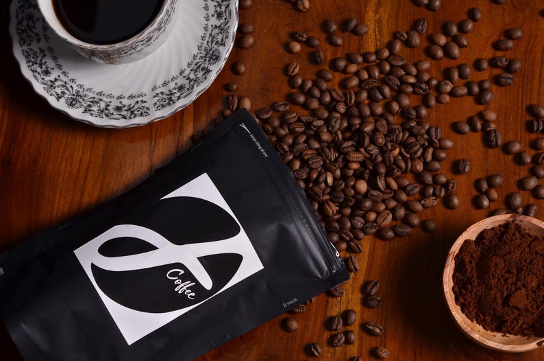 K&ouml;nnte beinhalten: Eine schwarze Kaffeet&uuml;te mit einem wei&szlig;en quadratischen Etikett, das einen stilisierten Buchstaben S und den Text Coffee zeigt, umgeben von verstreuten ger&ouml;steten Kaffeebohnen, einer wei&szlig;en Tasse mit schwarzem Blumenmuster und einem kleinen Holzsch&auml;lchen.