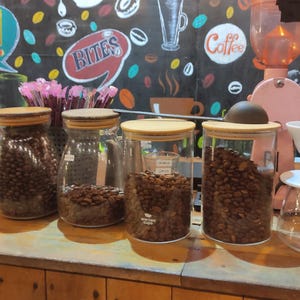 K&ouml;nnte beinhalten: Vier klare Glasbeh&auml;lter mit Holzdeckeln gef&uuml;llt mit ger&ouml;steten Kaffeebohnen, auf einer Holztheke neben einem wei&szlig;en Keramik-Kaffeefilter und einer rosa Kaffeem&uuml;hle, vor einer schwarzen Wand mit buntem, kaffeebezogenem Text platziert.