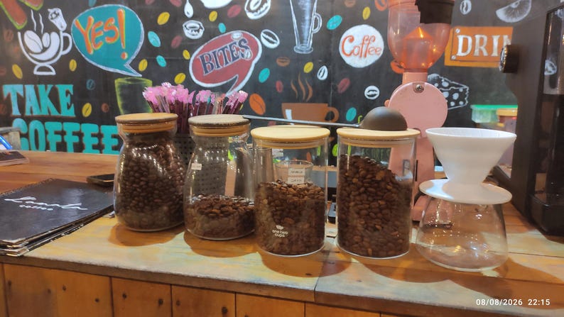K&ouml;nnte beinhalten: Vier klare Glasbeh&auml;lter mit Holzdeckeln gef&uuml;llt mit ger&ouml;steten Kaffeebohnen, auf einer Holztheke neben einem wei&szlig;en Keramik-Kaffeefilter und einer rosa Kaffeem&uuml;hle, vor einer schwarzen Wand mit buntem, kaffeebezogenem Text platziert.