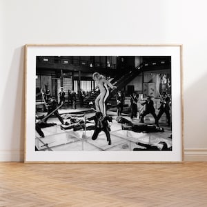 May include: A black and white framed print depicting a cinematic action scene with a central figure standing on another person's shoulders amidst a group of individuals in suits holding swords on a glass floor within a large interior space.