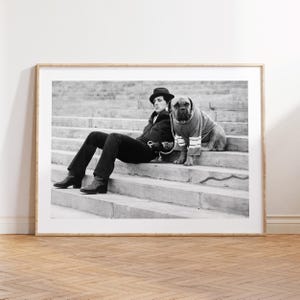 Może przedstawiać: Czarno-biała fotografia przedstawiająca osobę w ciemnej kurtce, spodniach, butach i kapeluszu typu fedora, siedzącą na kamiennych schodach obok dużego psa w swetrze w paski. Pies znajduje się tuż obok osoby na schodach w tej czarno-białej kompozycji.