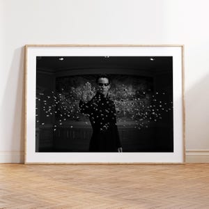 May include: A framed black and white photographic print featuring a person in a dark coat and sunglasses with one hand raised, surrounded by a multitude of suspended metallic spheres floating in the air within a dimly lit interior space.