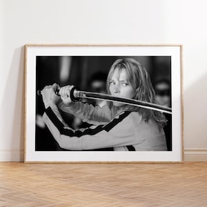 May include: A black and white framed print featuring a person with shoulder-length hair holding a katana sword horizontally in front of their face. The person wears a long-sleeved jacket with dark stripes along the arms, captured in a cinematic pose.