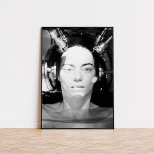 May include: A black and white framed print featuring a close-up portrait of a person with a neutral expression. The person is positioned within a circular metallic apparatus with multiple cylindrical components extending from the top and sides.