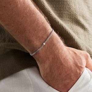 Può includere: Un bracciale a catena color argento con una singola piccola pietra rotonda e trasparente incastonata in una montatura a lunetta. Il bracciale è composto da sottili maglie metalliche intrecciate che formano un cinturino delicato.