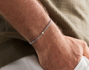Pode incluir: Uma pulseira de corrente em tom prateado com uma pequena pedra redonda e transparente cravada num engaste. A pulseira é composta por elos metálicos finos e entrelaçados que formam uma banda delicada e contínua à volta do pulso.