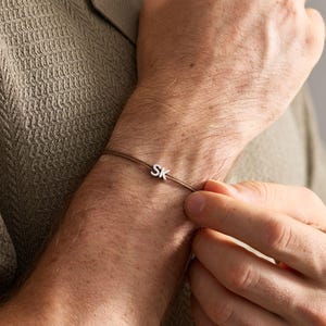 May include: A thin brown cord bracelet featuring a small, silver-toned metal charm with the letters SK engraved on it. The bracelet is worn around the wrist, showcasing the minimalist design of the cord and the metallic letter charm.