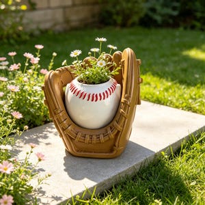 May include: A tan baseball glove-shaped planter holding a white baseball-shaped pot with red stitching. Small white daisy flowers with yellow centers are planted inside the baseball pot, creating a decorative display for a garden setting.