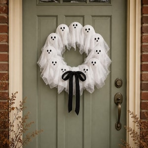 May include: A circular wreath composed of multiple small white ghost figures made of sheer fabric with black felt eyes and mouths. The ghosts are arranged in a ring, and a large black fabric ribbon is tied in a bow at the bottom centre.