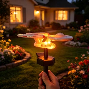Peut inclure: Une lampe de jardin en forme de vaisseau spatial blanc et gris avec l'inscription NCC-1701 sur le côté. Le modèle est monté sur un piquet noir et émet une lueur orange chaude depuis la section centrale, éclairant la structure dans l'obscurité.