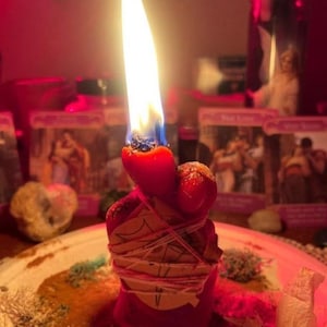 K&ouml;nnte beinhalten: Eine rote Stumpenkerze mit heller Flamme, umwickelt mit hellem Garn und einem kleinen St&uuml;ck Papier mit einem handgezeichneten Symbol. Die Kerze steht auf einem Teller, der mit verstreuten getrockneten Kr&auml;utern und Pflanzenteilen bedeckt ist.