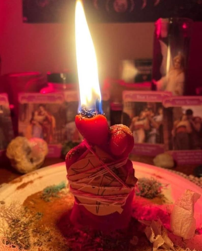 K&ouml;nnte beinhalten: Eine rote Stumpenkerze mit heller Flamme, umwickelt mit hellem Garn und einem kleinen St&uuml;ck Papier mit einem handgezeichneten Symbol. Die Kerze steht auf einem Teller, der mit verstreuten getrockneten Kr&auml;utern und Pflanzenteilen bedeckt ist.
